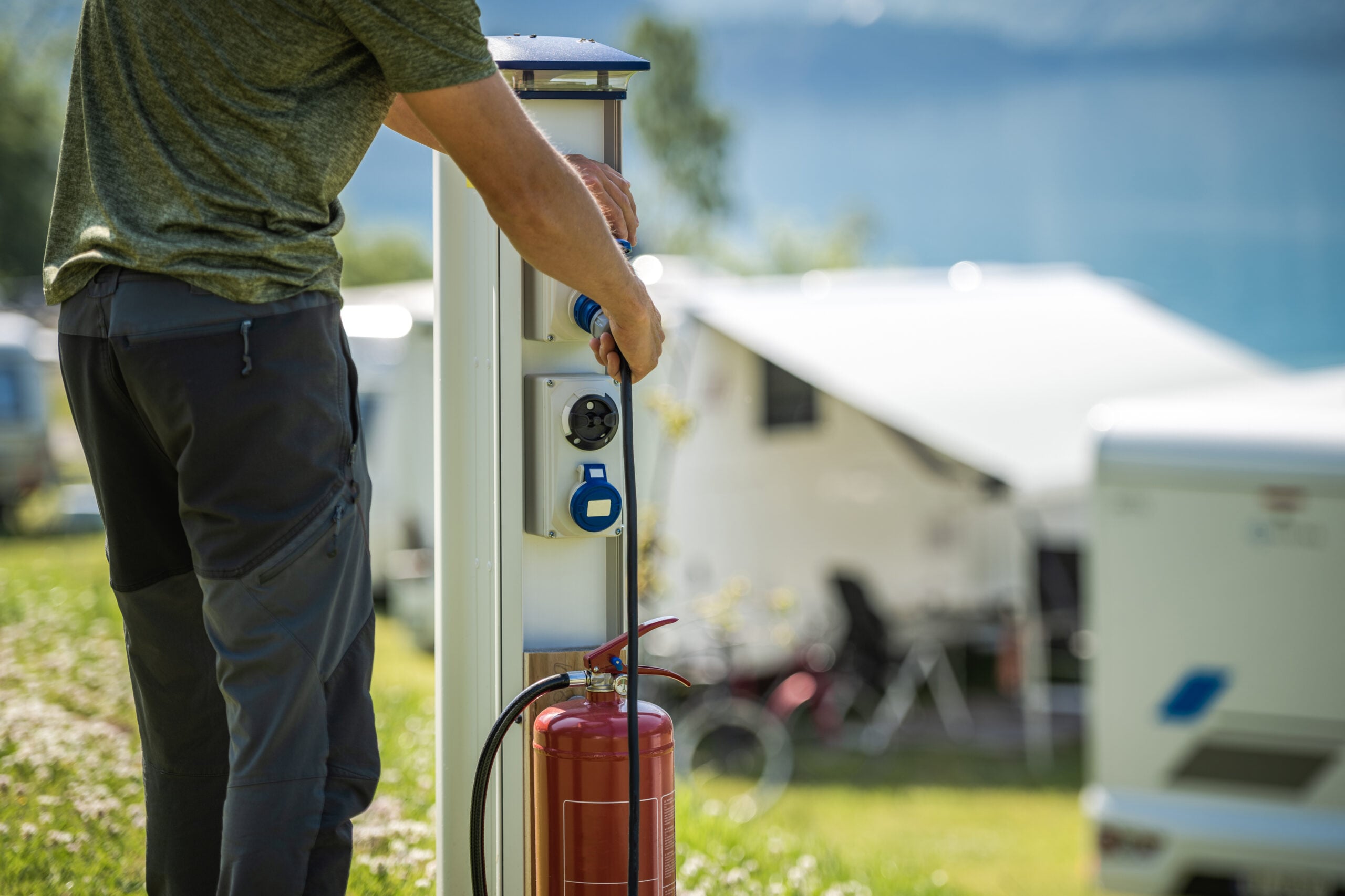 A person connects a cable and plug to an outdoor electrical hookup post at a campsite, with RVs and greenery visible in the background.