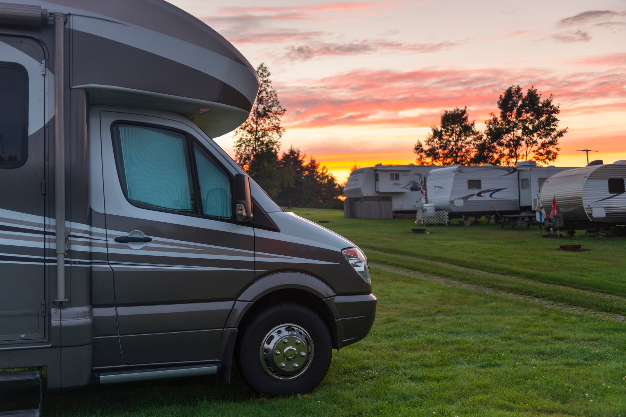 A gray RV is parked on green grass at a campground, with more RVs in the background. Trees and a colorful sunset sky with orange and pink clouds complete the peaceful outdoor scene.