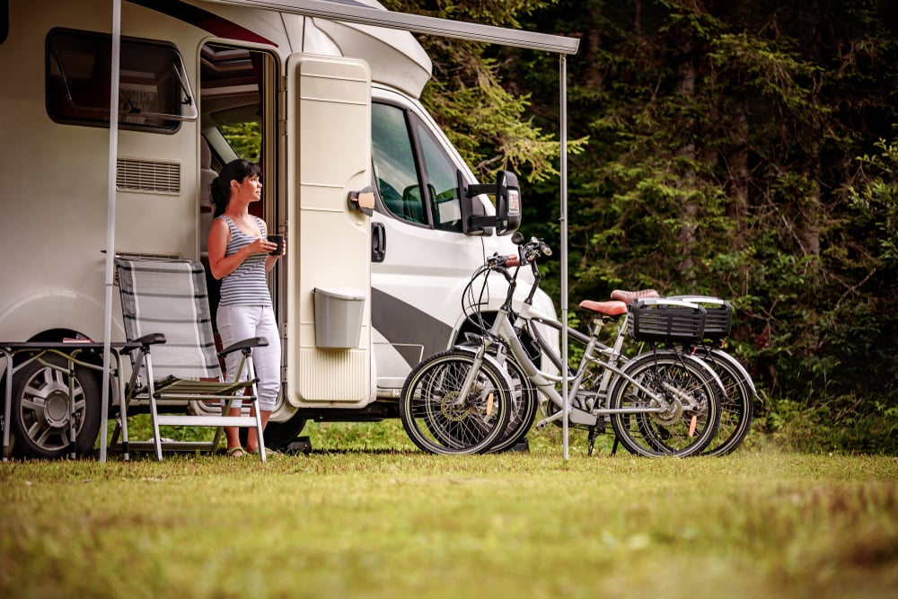 A woman stands by the open door of a camper van, holding a cup. Two bicycles are parked nearby on the grass. There are camping chairs outside and trees in the background.