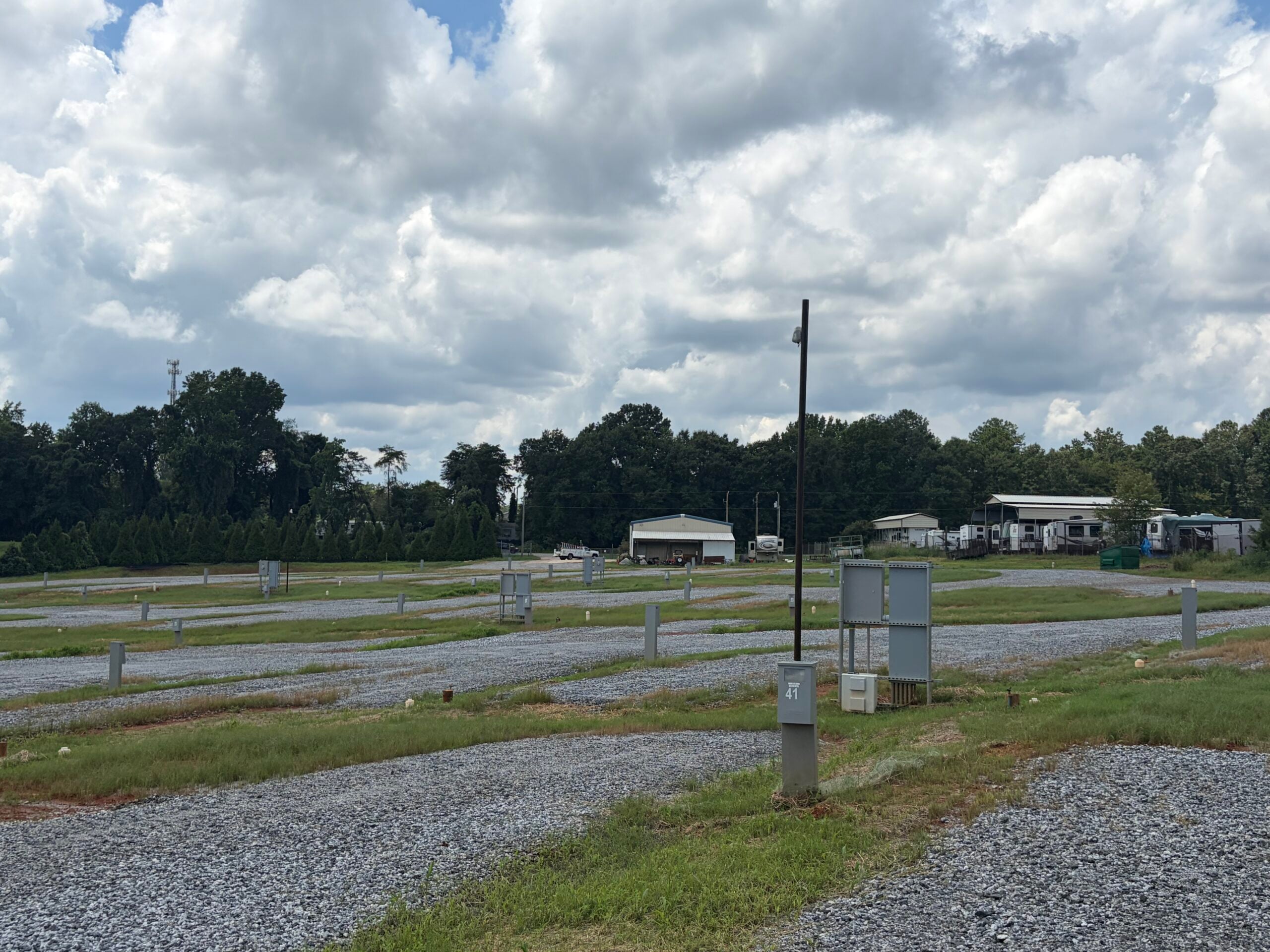 A mostly empty RV park with gravel parking spaces, utility hookups, and a few RVs and buildings in the background. Trees line the edge of the park under a partly cloudy sky.